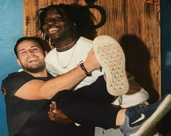A smiling man with a beard lifts another smiling man playfully in his arms. The lifted man wears a white shirt, jeans, and sneakers, while the man lifting him wears a dark shirt. They are in front of a wooden door with a blue wall.