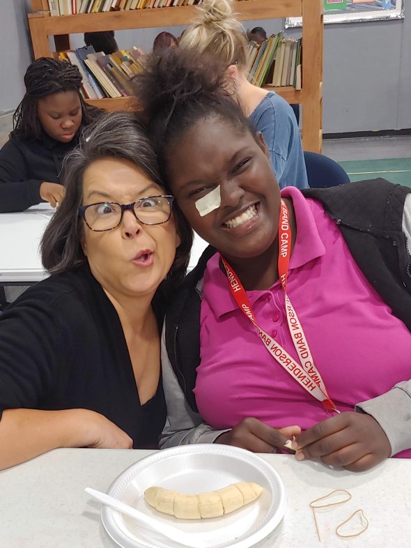 A woman with glasses playfully makes a funny face next to a smiling girl wearing a pink shirt and a red lanyard. They sit at a table with half a banana on a plate. Bookshelves are visible in the background.
