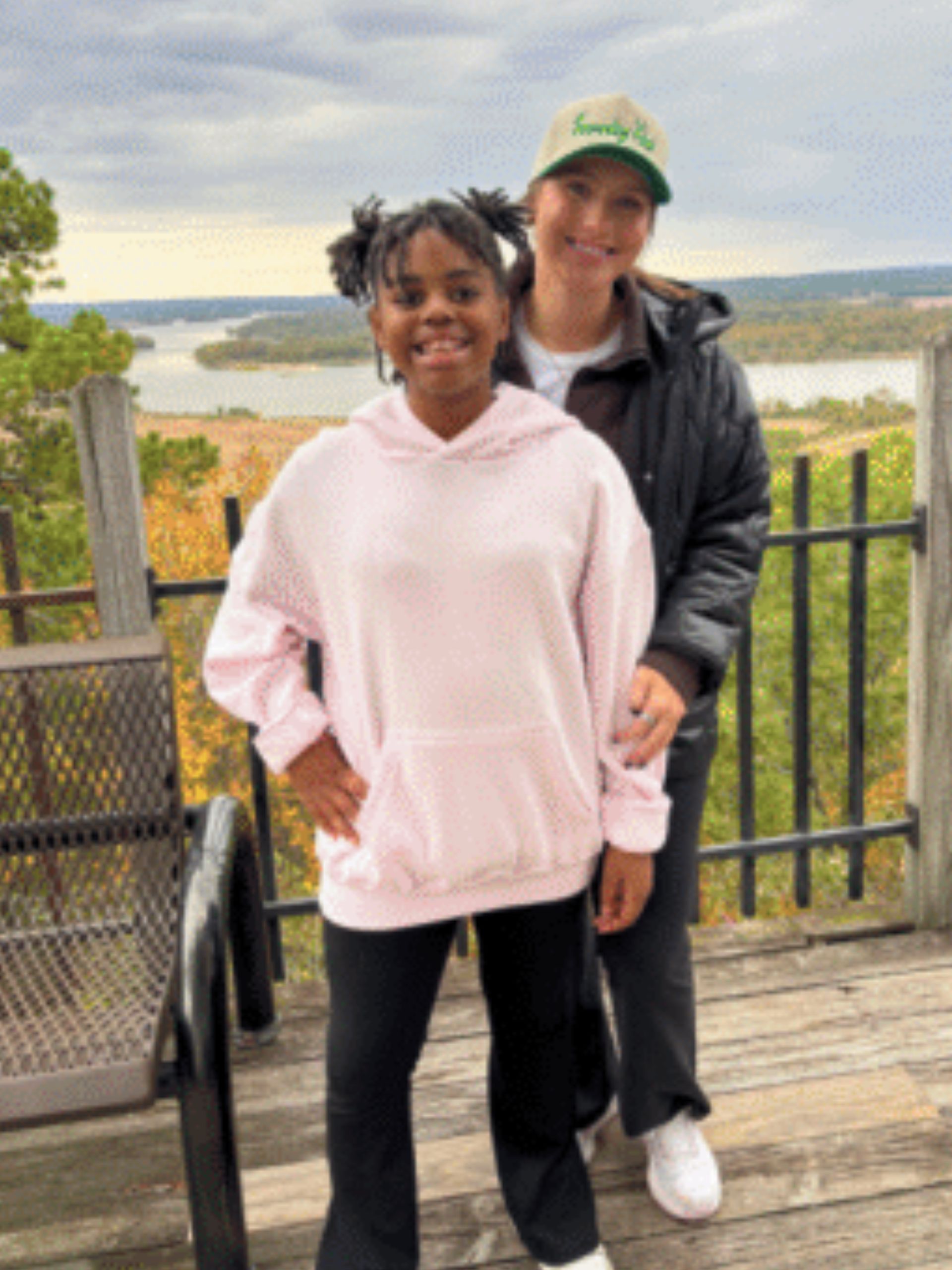 Two people stand smiling on a scenic overlook with trees, a river, and cloudy sky in the background. One wears a pink hoodie, the other a black jacket and green cap. A metal bench and railing are beside them.