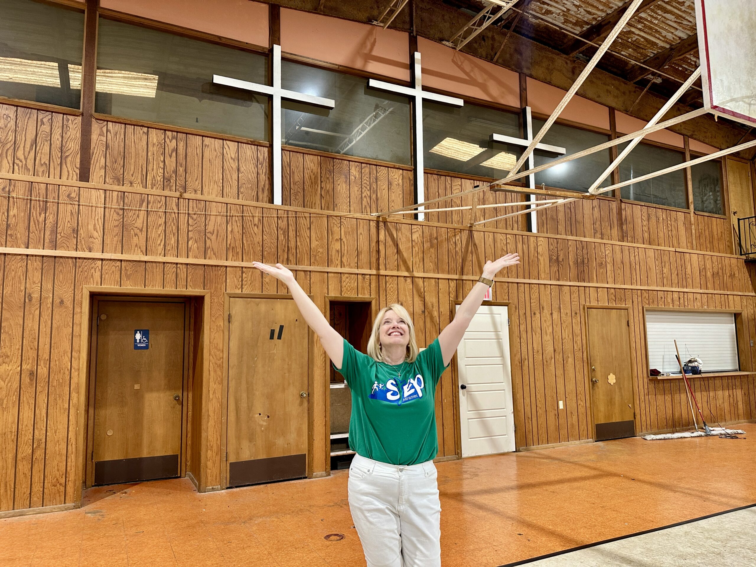 A smiling woman in a green shirt stands with arms raised in a gym with wood-paneled walls and three large white crosses above her. The space has doors, a mop, and a restroom sign visible.