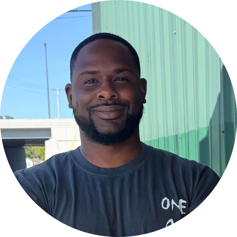 A man with short hair and a beard smiles while standing outside near a green corrugated metal wall, wearing a dark t-shirt with white text.